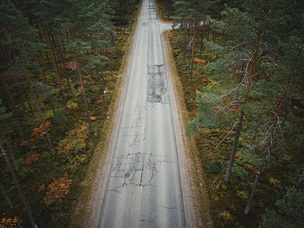 Suomalainen metsätie kiemurtelee tiheän mäntyjen keskellä, syksyiset värit ja kulunut asfaltti näkyvissä ylhäältä